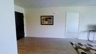 This single-floor residence offers a clean, modern design with an open and airy layout. The living area is finished with smooth wood-look flooring that extends throughout the space, complemented by crisp white walls and a neutral color palette that creates a bright, welcoming atmosphere. A distinctive architectural feature is the framed pass-through window built into the wall between the living area and the kitchen, providing visual connectivity between the two spaces while maintaining a sense of definition. Through the pass-through, the kitchen is visible and equipped with stainless steel appliances, including a microwave and a full-size refrigerator, set against clean cabinetry. The living area benefits from recessed lighting overhead, contributing to a well-lit interior environment. A doorway on the right side of the living area leads to an additional room, suggesting a functional multi-room floor plan. The overall design aesthetic leans contemporary and minimalist, with thoughtful architectural details that enhance both the practicality and visual appeal of the space. The single-level layout ensures ease of accessibility and a cohesive flow from room to room.