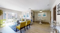 This single-floor residence offers a well-organized open-concept layout connecting the living, dining, and kitchen areas. The living room is bright and inviting, featuring light wood-style flooring, neutral warm-gray walls, and a sliding glass door that opens to a private balcony with outdoor views of surrounding greenery. The space accommodates comfortable seating and is enhanced by framed wall art in cool blue and green tones.

Adjacent to the living area, the dining space features a round white pedestal table paired with sleek acrylic and chrome chairs, lit by a flush ceiling fixture. Two framed coastal-themed prints adorn the wall, reinforcing the clean, contemporary aesthetic. A doorway nearby provides a glimpse into a well-appointed bathroom with vanity lighting.

The galley-style kitchen runs along a single corridor with white cabinetry on both sides, a mosaic tile backsplash, dual-basin sink, and black appliances including a range and refrigerator. A walk-in closet with built-in shelving is accessible directly from the kitchen. The bathroom includes a full soaking tub with white tile surround. Throughout the residence, light flooring and neutral walls create a cohesive, modern ambiance.