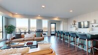 Located at 730 1/2 N First St in Minneapolis, MN, this contemporary communal living space offers a thoughtfully designed open-plan layout that maximizes both function and aesthetic appeal. The single-floor space is anchored by an expansive living area furnished with rich tan leather sofas, a glass coffee table, and layered area rugs atop warm, dark hardwood flooring that runs throughout.

A standout feature is the striking bar and lounge counter finished in a teal-painted base topped with dark, veined granite, accompanied by a full row of navy blue upholstered barstools. Behind the bar, a shiplap-style accent wall houses open shelving, a television, and subtle decorative lighting, creating an inviting focal point.

Floor-to-ceiling windows wrap around the space, flooding the interior with abundant natural light and offering sweeping panoramic views of the Minneapolis skyline and surrounding treetops. A glazed door leads to an exterior balcony equipped with high-top bistro tables and stools, extending the social atmosphere outdoors.

A dedicated lounge nook near the windows features additional high-top tables and a curated gallery wall of framed artwork, adding a creative and personalized touch. Recessed ceiling lighting and in-ceiling speakers complete the modern, polished ambiance, making this space ideal for gathering, entertaining, and relaxing.