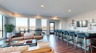 Located at 730 1/2 N First St in Minneapolis, MN, this contemporary communal living space offers a thoughtfully designed open-plan layout that maximizes both function and aesthetic appeal. The single-floor space is anchored by an expansive living area furnished with rich tan leather sofas, a glass coffee table, and layered area rugs atop warm, dark hardwood flooring that runs throughout.

A standout feature is the striking bar and lounge counter finished in a teal-painted base topped with dark, veined granite, accompanied by a full row of navy blue upholstered barstools. Behind the bar, a shiplap-style accent wall houses open shelving, a television, and subtle decorative lighting, creating an inviting focal point.

Floor-to-ceiling windows wrap around the space, flooding the interior with abundant natural light and offering sweeping panoramic views of the Minneapolis skyline and surrounding treetops. A glazed door leads to an exterior balcony equipped with high-top bistro tables and stools, extending the social atmosphere outdoors.

A dedicated lounge nook near the windows features additional high-top tables and a curated gallery wall of framed artwork, adding a creative and personalized touch. Recessed ceiling lighting and in-ceiling speakers complete the modern, polished ambiance, making this space ideal for gathering, entertaining, and relaxing.