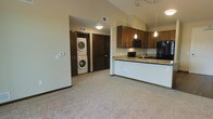 This single-floor unit showcases an open-concept design that seamlessly connects the living area and kitchen. Vaulted ceilings enhance the sense of space throughout, while neutral carpet flooring in the main living area adds warmth and comfort underfoot.

The kitchen is outfitted with rich, medium-toned wood cabinetry and granite countertops featuring warm tonal variation. Black appliances, including a microwave and refrigerator, complement the cabinetry, while a peninsula with a built-in sink provides additional prep space and a natural divide between the kitchen and living area. Pendant lighting hangs above the peninsula, and a track lighting system overhead ensures ample illumination.

A dedicated laundry area is conveniently integrated into the unit, housing a stacked washer and dryer. Dark-stained interior doors add a cohesive design element throughout the space, contrasting neatly with the light-toned walls. A visible thermostat and ceiling vent reflect practical, functional detailing. The overall ambiance is clean and contemporary, with a neutral palette that provides a versatile backdrop.
