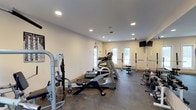This fitness room offers a comprehensive workout environment designed to accommodate a variety of exercise routines. The space features durable rubber-style flooring suited for heavy equipment use, and is illuminated by recessed ceiling lights that provide bright, even coverage throughout. Natural light filters in through windows near the entrance doors, contributing to an open and energizing atmosphere.

The room is equipped with a treadmill and additional cardio machines, making it suitable for sustained aerobic workouts. On one side, a cable machine and suspension training system offer versatile strength and functional training options. Along the opposite wall, a rack of dumbbells provides a range of free weight options, complemented by a squat rack with a barbell for compound strength training. A wall-mounted flat-screen television adds a convenient entertainment element for those engaging in longer workout sessions.

The neutral white walls and clean layout keep the space feeling organized and functional. Overall, the room is thoughtfully arranged to maximize usability and provide residents or users with a well-rounded fitness facility.