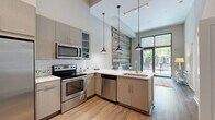This thoughtfully designed one-bedroom, one-bathroom unit offers a contemporary living experience across a single floor. Soaring ceilings and warm hardwood-style flooring establish an open, airy ambiance throughout the space. The kitchen is a standout feature, showcasing a suite of stainless steel appliances — including a range, microwave, dishwasher, and refrigerator — complemented by sleek gray cabinetry, white subway tile backsplash, and light quartz countertops. Three industrial-style pendant lights hang above the kitchen peninsula, while a striking reclaimed wood accent wall adds rustic character. Floor-to-ceiling windows and an adjacent outdoor balcony flood the area with natural light and provide views of the surrounding streetscape and greenery. The bedroom is comfortably sized, featuring large windows that frame tree-lined street views, soaring ceiling height, and direct access to a dedicated closet and the full bathroom. The walk-in closet is outfitted with a comprehensive wire shelving and storage system. The bathroom offers large-format tile flooring, a double-door glass shower, and a gray vanity with ample storage, further enhanced by a sizable window that brings in natural light. The overall design balances modern finishes with warm, inviting details.