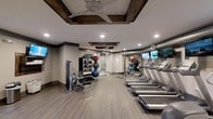 This fitness center combines modern functionality with distinctive architectural character. The ceiling is a standout feature, showcasing rich, dark wood beams set within coffered panels, complemented by a ceiling fan and recessed lighting that together provide both style and effective illumination throughout the space.

The flooring consists of durable, wood-look planks in a warm neutral tone, lending a clean and polished appearance underfoot. Along one side of the room, a row of treadmills is neatly arranged, each equipped with individual screens for an engaging workout experience. Wall-mounted televisions are positioned for easy viewing from multiple points in the room.

On the opposite side, a strength and functional training area houses a cable machine, a weight bench, and a well-organized rack stocked with medicine balls and stability balls in various sizes. A large mirror spans the wall in this area, enhancing the sense of depth and allowing users to monitor their form. Exposed ductwork along the upper walls adds an industrial accent that complements the rustic beam detailing, resulting in a space that is both practical and visually engaging.
