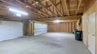 This single-story garage offers a generous open floor plan with a poured concrete floor, providing a clean and durable surface suited for a wide range of uses, from vehicle storage to workshop activities. The interior walls display exposed wood stud framing, lending the space a raw, utilitarian character while leaving room for customization such as insulation, drywall, or shelving installation. Overhead, an open truss ceiling with visible wooden rafters contributes to a notably tall clearance, accommodating larger vehicles or overhead storage solutions. A sectional garage door provides convenient access, complemented by an automatic door opener mounted to the ceiling track. Fluorescent overhead lighting fixtures illuminate the interior, ensuring adequate visibility throughout the space. The expansive, unobstructed floor area offers considerable flexibility, making it well-suited for use as a multi-car garage, storage facility, or hobby and workshop space. The straightforward, no-frills construction emphasizes functionality, and the existing wood-framed walls provide a solid foundation for any future finishing or improvement projects tailored to the occupant's needs.