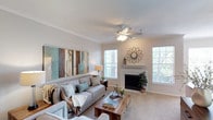 This single-floor residence in Lewisville offers a thoughtfully designed living space that balances comfort and style. The living room serves as the heart of the home, showcasing a classic fireplace with a white-painted mantle and decorative accents that add a warm, inviting focal point to the room. Crown molding lines the ceiling, lending a refined architectural detail that elevates the overall aesthetic. A ceiling fan with integrated lighting ensures year-round comfort, while multiple windows fitted with plantation-style shutters flood the space with generous natural light and provide a view of the surrounding greenery outside. The neutral, light-toned walls and plush carpeted flooring create a calm and airy ambiance throughout. The layout is open and spacious, accommodating a generous seating arrangement alongside a wooden coffee table that grounds the room with a touch of natural texture. Tasteful wall art featuring muted tones of teal, cream, and wood complement the overall color palette, contributing to a cohesive and serene interior. This space is well-suited for both relaxed everyday living and comfortable entertaining.