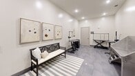 Located at 13415 Old Statesville Rd in Huntersville, NC, this dedicated pet grooming suite is a single-floor space designed to deliver a professional and welcoming experience for pets and their owners alike. The room is finished with crisp white walls and large-format grey tile flooring, creating a clean, modern aesthetic throughout.

The grooming area is equipped with multiple stainless steel bathing stations featuring retractable hose attachments and step-up access ramps, ensuring ease of use for pets of varying sizes. Complementing these are hydraulic lift grooming tables outfitted with adjustable restraint arms, providing a safe and functional workspace for professional grooming. A convenient storage and prep counter with a dark cabinetry base and light countertop offers ample workspace and organization.

Recessed ceiling lighting illuminates the space evenly, while higher ceilings lend an open, airy feel. A striped area rug and a cushioned wooden bench offer a comfortable waiting area, complemented by a curated selection of framed wall art and decorative greenery that softens the professional environment. Additional amenities include a small round side table and mounted accessory organizers. The suite is accessed through a private door, ensuring a focused and contained grooming environment ideal for a residential community amenity.