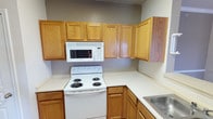 This single-floor residence offers a straightforward and functional layout designed for comfortable everyday living. The kitchen is well-equipped with warm oak cabinetry spanning both upper and lower sections, light-toned countertops, a built-in over-the-range microwave, a white electric coil range, and a double stainless steel sink. The L-shaped kitchen configuration provides ample counter and storage space, opening toward the main living area. The living area features neutral-toned carpet, crown molding, a ceiling fan, and large sliding glass doors dressed with vertical blinds that allow natural light to fill the space. Adjacent to the living area, a hallway leads to the bathroom, which includes a vanity with a sink, mirror, overhead wall cabinet for storage, and a toilet. The bedrooms are carpeted and outfitted with ceiling fans; one bedroom benefits from natural light through a pair of windows with blinds, while another features a more enclosed, private feel. Throughout the home, walls are painted in soft neutral tones, and crown molding accents several areas, adding a refined touch. The combination of hardwood-style flooring in the kitchen and transitional areas with carpet in the living and sleeping spaces creates a cohesive and practical interior.