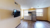 This single-floor unit is organized into a compact yet functional layout encompassing a combined living and kitchen area, two separate rooms, and a full bathroom. The living area features wood-look plank flooring and warm yellow-toned walls, with a textured ceiling overhead. A wall-mounted flat-screen TV is positioned near the entry, and the open sightlines to the kitchen make the shared space feel cohesive and practical.

The kitchen is outfitted with white shaker-style cabinetry — both upper and lower — providing ample storage. Stainless steel appliances include a full-size refrigerator, a built-in microwave, and a cooktop. A stainless steel sink with additional cabinetry is positioned along a separate wall, and multiple windows allow natural light to fill the area while offering exterior views.

The two additional rooms are carpeted with a bold geometric-patterned carpet in gold and blue tones, contrasting with the lighter walls. One room includes a closet, adding storage utility. Both rooms are well-proportioned with natural light from single windows fitted with blinds.

The bathroom features a dark wood-toned vanity with a white countertop, a framed mirror, chrome fixtures, and wood-look flooring that carries a consistent material palette throughout the unit.