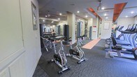 This fitness center offers a thoughtfully organized single-floor layout designed to accommodate a wide range of workout routines. The space is divided into distinct functional zones, with a cardio area featuring a row of stationary cycling bikes positioned along rubberized flooring for comfort and safety during high-intensity sessions. Adjacent to the cardio zone, a dedicated strength training area houses multiple cable and resistance machines, providing users with versatile options for targeted muscle training. A free weights section, complete with a full dumbbell rack, is reflected in floor-to-ceiling mirrors that line one wall, enhancing the sense of space and allowing users to monitor their form. The flooring transitions between carpet, rubber mat, and a warm-toned runner, clearly delineating each workout zone. Recessed lighting throughout keeps the space well-lit, while ceiling-mounted pendant fans provide airflow during intense workouts. Structural columns are integrated seamlessly into the layout, and informational charts are posted on the walls for user reference. The overall ambiance is functional and energizing, offering a clean, well-maintained environment suited for a variety of fitness goals.