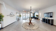 Michigan Street Commons presents a thoughtfully designed single-floor communal space that blends contemporary aesthetics with functional comfort. The spacious living room serves as the heart of the space, anchored by a striking round pedestal table atop a patterned area rug and illuminated by a sleek multi-arm chandelier. Natural light pours in through large glass double doors, lending the area an open and airy atmosphere. A bold geometric accent wall in deep blue and gold tones creates a dramatic focal point, complemented by a seating nook with dark upholstered chairs. A vibrant urban mural adds a creative, energetic element to the room, while a wall-mounted TV and modern console table provide additional utility. The hallway extends the design language of the living room with light-toned luxury vinyl flooring, crisp white walls, and curated wall art. Tufted benches offer seating along the corridor, which is further dressed with potted greenery and the prominently displayed Michigan Street Commons branding. Elevator access and additional doors along the hallway ensure seamless connectivity throughout. Together, the two areas form a cohesive, design-forward communal environment that is both welcoming and functional.
