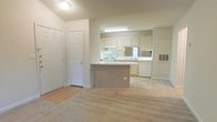 This single-floor space features an open-concept design that integrates the living area and kitchen into one cohesive layout. The living area is finished with light-colored carpet that extends throughout, creating a neutral and understated foundation. Near the entrance, hardwood-style flooring provides a practical transition zone, accompanied by a closet with a paneled door offering convenient storage.

The kitchen is positioned toward the rear of the open space and is defined by a curved peninsula that serves as both a workspace and a visual divider. White cabinetry lines the walls, providing ample storage, while white appliances — including a range and dishwasher — maintain a clean, uniform aesthetic. A window above the sink allows natural light to brighten the kitchen area. Upper cabinets with a mirrored backsplash add a sense of depth to the space. A wall-mounted utility panel is also visible near the kitchen, adding to the functional elements of the layout. The overall palette of whites and light neutrals throughout gives the space a bright, open, and airy ambiance.