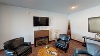 This single-floor space is thoughtfully designed to serve as a multifunctional gathering and community area, combining comfortable lounge seating, formal dining, casual café-style seating, and a fully equipped kitchen under one roof.

The lounge area anchors one end of the space, centered around a fireplace topped with a wall-mounted flat-screen television. Rich, dark carpeting and leather armchairs arranged around a wooden coffee table create a warm, relaxed atmosphere. Decorative touches, including a boat-shaped bookshelf and a console table with a lantern, lend a rustic, lodge-inspired character. A pass-through window connects this area to an adjacent room, enhancing the open flow of the space.

A large oval dining table with ample seating occupies the central zone, suitable for group meetings or communal meals. Nearby, a café-style high-top table and chairs are positioned beside large windows that offer views of the surrounding outdoor area, filling the space with natural light.

The kitchen is generously sized and well-appointed, featuring dark wood cabinetry, granite countertops, stainless steel appliances including a refrigerator, range, dishwasher, and microwave, and a spacious island with bar seating. Hardwood-style flooring throughout the kitchen zone contrasts with the carpeted lounge, creating a clear and functional delineation between areas.