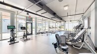 Located at 160 Morgan St in Jersey City, NJ, this fitness center offers a generously sized, single-floor facility designed to accommodate a comprehensive range of workout routines. The space is organized into distinct functional zones, each outfitted with professional-grade equipment.

The cardio area is a standout feature, lined with floor-to-ceiling windows that flood the room with natural light and offer views of the surrounding outdoor greenery and urban streetscape. Treadmills, elliptical trainers, stair climbers, and rowing machines are arranged in orderly rows, providing ample space between stations. Large mirrors along the walls further enhance the sense of openness.

A dedicated strength and free weights section features adjustable benches, a full dumbbell rack, cable machines, and multi-use weight systems, all set against a durable rubber flooring surface ideal for heavy lifting. A wall-mounted flat-screen TV provides entertainment in this area.

The spin studio corner houses several Peloton-style stationary bikes alongside step platforms and stability balls, offering versatility for cycling and group fitness-style workouts. Exposed ductwork and industrial-style ceilings contribute to the modern, functional aesthetic throughout. The facility also includes restrooms and a water fountain, ensuring convenience for all users.