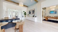 Located in Santa Rosa Beach, Florida, this beautifully appointed clubhouse blends coastal charm with modern design. The single-floor layout features soaring ceilings accented by exposed natural wood beams, lending warmth and character throughout the space. Light-washed hardwood flooring runs seamlessly across the open interior, reflecting the abundant natural light that pours in through oversized French doors and transom windows facing an outdoor pool area.

The main entrance welcomes visitors with a striking round pedestal table serving as a focal centerpiece, topped with curated décor including tall ceramic vases and fresh floral arrangements. A sky-blue beadboard accent wall adds a distinctly coastal touch, complemented by crisp white shiplap paneling in the lounge area. Comfortable seating includes a tufted gray sofa adorned with ocean-toned throw pillows, positioned beneath a large-format seascape photograph that anchors the space.

A built-in white sideboard provides functional storage and display space, while a small beverage station offers added convenience. Woven rattan chairs and round café tables create casual seating vignettes throughout. Additional rooms, accessible through interior French doors, appear to include a fitness area. The overall ambiance is airy, welcoming, and thoughtfully designed to reflect the relaxed, upscale lifestyle of the Florida Gulf Coast.