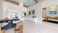 Located in Santa Rosa Beach, Florida, this beautifully appointed clubhouse blends coastal charm with modern design. The single-floor layout features soaring ceilings accented by exposed natural wood beams, lending warmth and character throughout the space. Light-washed hardwood flooring runs seamlessly across the open interior, reflecting the abundant natural light that pours in through oversized French doors and transom windows facing an outdoor pool area.

The main entrance welcomes visitors with a striking round pedestal table serving as a focal centerpiece, topped with curated décor including tall ceramic vases and fresh floral arrangements. A sky-blue beadboard accent wall adds a distinctly coastal touch, complemented by crisp white shiplap paneling in the lounge area. Comfortable seating includes a tufted gray sofa adorned with ocean-toned throw pillows, positioned beneath a large-format seascape photograph that anchors the space.

A built-in white sideboard provides functional storage and display space, while a small beverage station offers added convenience. Woven rattan chairs and round café tables create casual seating vignettes throughout. Additional rooms, accessible through interior French doors, appear to include a fitness area. The overall ambiance is airy, welcoming, and thoughtfully designed to reflect the relaxed, upscale lifestyle of the Florida Gulf Coast.
