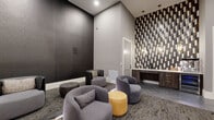 This single-floor screening and lounge space is designed with a refined, contemporary aesthetic that balances comfort with visual impact. The room's most striking architectural feature is its dramatically tall ceiling, which amplifies the sense of openness and grandeur. A floor-to-ceiling mosaic accent wall composed of dark, gold, and metallic tile pieces serves as a bold focal point, illuminated by warm pendant lighting that adds an inviting glow to the space.

The lounge area is furnished with a curated mix of rounded, upholstered armchairs in charcoal and gray tones, complemented by mustard-yellow ottomans and low circular side tables. The seating is arranged on a patterned, textured area rug that adds depth and warmth to the space. On the opposite end of the room, a large projection screen is mounted against a dark feature wall accented by vertical wood slat paneling, creating an intimate cinematic atmosphere.

A built-in refreshment counter with dark wood cabinetry, a beverage cooler, and a countertop for snacks and drinks provides added convenience. A gold-framed bar cart offers additional display space. Hexagonal tile flooring transitions into the carpeted lounge zone, and a dark paneled exit door is accessible at the rear of the room.