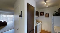 This single-floor space is organized around a practical layout that connects a hallway corridor to a compact kitchen area. The kitchen is equipped with a white electric coil-burner range and oven, complemented by dark wood cabinetry overhead that provides ample storage. Light-toned countertops offer a contrast to the darker cabinet finish, and a range hood is mounted above the stove. Track lighting runs along the kitchen ceiling, providing directed illumination throughout the cooking area. A full-height dark wood door, likely a pantry or closet, serves as a prominent dividing element between the hallway and kitchen space, adding both functionality and visual weight to the transition zone. The hallway features white walls adorned with a small wall-mounted decorative sconce, adding a subtle decorative touch. Framed artwork hangs on the kitchen wall, giving the space a personalized feel. The flooring transitions from hard surface in the kitchen to carpet in the adjacent hallway and living areas, visible in the background. Natural light appears to filter in from a nearby window, contributing to the overall brightness of the space.