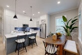 Located in Fort Mill, SC, this single-story, 2-bedroom, 2-bathroom home offers a thoughtfully designed layout that balances comfort and functionality. The open-concept kitchen and dining area serves as the heart of the home, showcasing white shaker cabinetry extending to the ceiling, a striking blue subway tile backsplash, stainless steel appliances, a two-tone island with seating, and light hardwood-style flooring throughout. Pendant lighting and recessed fixtures illuminate the space, while a large window in the dining nook invites natural light and outdoor views. The living room features a wall of windows, flooding the area with natural light and providing a bright, welcoming atmosphere complemented by crown molding and a ceiling fan. A dedicated office offers a quiet retreat with carpeted flooring and a ceiling fan. The generously sized primary bedroom includes a closet with double arched-panel doors. The second bedroom is well-lit with multiple windows and plush carpeting. Both bathrooms are finished with large-format tile flooring and surrounds, white vanities, and clean, neutral palettes. The overall ambiance throughout the home is contemporary yet warm, with a cohesive neutral color scheme and modern finishes.
