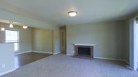 This single-story space presents a straightforward and functional layout centered around a comfortable living area. The main room is anchored by a classic brick fireplace with a white painted mantle, serving as a natural focal point. The fireplace's traditional red brick surround adds a warm, timeless character to the space. Neutral sage-green walls complement the light carpeted flooring throughout the living area, creating a calm and understated ambiance. A flush-mount ceiling light provides overhead illumination, contributing to the room's practical and clean aesthetic. The flooring transitions from carpet in the living area to hardwood or laminate in an adjacent section, visible toward the back of the space, adding subtle variety. A hallway visible from the main room connects to additional areas of the home, suggesting a practical flow between spaces. White baseboards run along the perimeter of the walls, providing a clean contrast to the neutral wall color. The overall design is modest and move-in ready, offering a blank canvas that lends itself well to a variety of interior styles and furnishing choices.