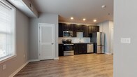 This well-appointed single-floor unit offers a thoughtfully designed layout that balances functionality with modern style. The kitchen serves as a standout feature, showcasing rich dark cabinetry that extends to the ceiling, a classic white subway tile backsplash, granite countertops, and a full suite of stainless steel appliances including a range, over-the-range microwave, dishwasher, and French door refrigerator. Warm wood-style luxury vinyl plank flooring runs throughout the main living areas, complementing the neutral gray wall tones and crisp white trim. Natural light filters into the open living and kitchen area through a large window fitted with blinds, creating a bright and inviting atmosphere. Recessed lighting provides additional illumination throughout. The bathroom continues the design cohesion with dark cabinetry, granite countertops, a large mirror, and clean tile flooring. A dedicated in-unit laundry area with a stacked washer and dryer adds a practical convenience. The bedroom area offers a closet with a hanging rod, while louvered and panel-style doors add subtle architectural character. The overall layout, visible in the dollhouse view, is efficient and open, making excellent use of the available space.