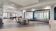 This dynamic amenity facility is thoughtfully designed to offer a range of social, recreational, and professional environments under one roof. The space is anchored by a generous game room featuring a full-size pool table, ping-pong table, and arcade machine, all set atop a combination of light hardwood and patterned flooring. Bold, colorful murals and vibrant wallpaper accent the walls, infusing energy into the space.

A comfortable lounge area adjoins the game room, furnished with a deep leather sectional, a patterned area rug, and multiple wall-mounted TVs — ideal for group gatherings or casual relaxation. Natural light streams in through large, dark-framed windows that frame views of the surrounding exterior.

A dedicated conference room features a long executive-style table surrounded by rolling chairs, statement pendant lighting, and a vivid patterned carpet, making it well-suited for meetings or events. A smaller breakout room with a round table and TV offers an intimate setting for smaller groups.

Glass interior windows and partitions create visual connectivity between zones, while a curved decorative railing near the entrance adds an architectural focal point. A dual water fountain set against a colorful mosaic tile wall provides a refined utilitarian detail. Throughout, the design balances bold creative expression with functional, well-appointed spaces.