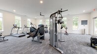 Located at 3506 Buster Lane in Indian Land, SC, this fitness center is designed to accommodate a wide range of workout styles within a single, open-plan floor. The space is generously proportioned, offering ample room for users to move freely between equipment stations without feeling crowded.

The interior features neutral white walls and textured gray carpet tiles throughout, contributing to a clean, modern aesthetic. Recessed lighting and multiple ceiling fans are evenly distributed across the ceiling, ensuring consistent illumination and airflow. Large, white-framed windows line the perimeter walls on multiple sides, flooding the space with natural light and offering views of the surrounding greenery.

The strength training area is well-appointed with True-brand cable machines, a power rack with an Olympic barbell and weight plates, a dumbbell rack with a full range of free weights, and a flat bench. A wall-length mirror in this section supports form monitoring during lifts. The cardio section includes treadmills, ellipticals, stair climbers, a rowing machine, and stationary spin bikes. Functional training is also supported through TRX suspension straps, a tire, and a multi-purpose functional trainer rack with accessories. A wall-mounted flat-screen television is positioned for visibility across the space.