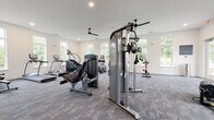 Located at 3506 Buster Lane in Indian Land, SC, this fitness center is designed to accommodate a wide range of workout styles within a single, open-plan floor. The space is generously proportioned, offering ample room for users to move freely between equipment stations without feeling crowded.

The interior features neutral white walls and textured gray carpet tiles throughout, contributing to a clean, modern aesthetic. Recessed lighting and multiple ceiling fans are evenly distributed across the ceiling, ensuring consistent illumination and airflow. Large, white-framed windows line the perimeter walls on multiple sides, flooding the space with natural light and offering views of the surrounding greenery.

The strength training area is well-appointed with True-brand cable machines, a power rack with an Olympic barbell and weight plates, a dumbbell rack with a full range of free weights, and a flat bench. A wall-length mirror in this section supports form monitoring during lifts. The cardio section includes treadmills, ellipticals, stair climbers, a rowing machine, and stationary spin bikes. Functional training is also supported through TRX suspension straps, a tire, and a multi-purpose functional trainer rack with accessories. A wall-mounted flat-screen television is positioned for visibility across the space.