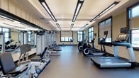 This fitness center offers a comprehensive workout environment designed to accommodate a wide range of exercise routines. The space is thoughtfully laid out, with cardio equipment — including treadmills and elliptical machines — arranged along one side, and a dedicated strength training area featuring cable machines, weight benches, and free weights on the other. Large windows line the walls, allowing natural light to fill the room and providing views of the greenery outside, creating an energizing atmosphere for workouts. Full-length mirrors along one wall enhance the sense of openness while also allowing users to monitor their form. The flooring consists of durable, dark rubberized material well-suited to high-traffic fitness use. Overhead, a coffered ceiling with modern linear LED lighting ensures the space is brightly and evenly illuminated throughout. The neutral wall tones and clean layout contribute to a focused, professional fitness environment. The single-floor facility is well-maintained and organized, making it accessible and functional for users of varying fitness levels.