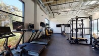 Located at 20060 Prairie St in the Chatsworth neighborhood of Los Angeles, this well-appointed fitness center offers a thoughtfully designed, single-floor workout facility suited for a wide range of training needs. The space features soaring ceilings with exposed ductwork and industrial-style ceiling fans, lending the gym a contemporary, open-air aesthetic. Durable rubber flooring covers the entire workout area, providing safety and comfort underfoot throughout.

The cardio section is equipped with treadmills, stationary bikes, and a stair climber, each paired with individual display screens for entertainment during workouts. Mounted flat-screen TVs are positioned throughout the space for additional viewing options. The strength training area offers an impressive array of equipment, including cable machines, multi-functional weight stations, a power rack, leg press, and a dumbbell rack, catering to both beginners and experienced athletes.

Large floor-to-ceiling windows and glass sliding doors flood the interior with abundant natural light while offering views of the surrounding outdoor areas, including what appears to be a pool deck. The open layout allows for generous spacing between equipment, ensuring a comfortable and safe training environment. The overall ambiance is clean, modern, and energizing, making this fitness center a well-rounded facility for daily exercise.