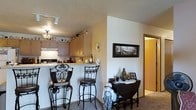 This single-floor residence offers an open, connected layout that blends the kitchen and living areas seamlessly. The kitchen features light wood cabinetry extending to the ceiling, providing ample storage space, and is equipped with a smooth countertop that transitions into a raised breakfast bar. The bar area is complemented by two ornate wrought-iron bar stools, adding a decorative yet functional touch to the space. Track lighting above the kitchen illuminates the workspace, while a Tiffany-style pendant light adds a warm, vintage charm to the area. A compact appliance setup, including a coffee maker and other countertop essentials, keeps the kitchen practical and organized. Adjacent to the kitchen, the dining nook features a dark wood drop-leaf table paired with a matching chair, accented by a lace table runner and decorative objects. A framed landscape photograph on the wall adds visual interest to the neutral-toned walls. The flooring transitions from tile in the kitchen to carpet in the living and dining areas, and a hallway visible in the background suggests additional rooms beyond, contributing to the functional flow of this cozy, well-appointed home.