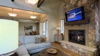 This single-story space showcases a warm and inviting design highlighted by a dramatic floor-to-ceiling stone fireplace, which serves as the focal point of the living area. A wall-mounted television is positioned above the fireplace, seamlessly integrating modern amenities with the rustic aesthetic. The living room features vaulted ceilings accented by exposed wood beams, adding architectural character and a sense of openness to the space. Light hardwood flooring runs throughout, complementing the neutral wall tones and contributing to a cohesive, natural palette. A comfortable sectional sofa with decorative pillows is arranged around a round coffee table, creating an ideal gathering area. An open passage connects the living space to a dedicated dining area, which features pendant and chandelier lighting, a dining table with upholstered chairs, and framed wall art. Large windows throughout allow natural light to fill both areas. A wooden sideboard in the living room provides functional storage, while a table lamp adds warm ambient lighting. The overall ambiance is rustic yet refined, balancing comfort with tasteful design details.