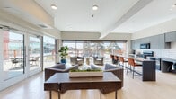Located in El Cerrito, California, this single-floor space is designed with a contemporary aesthetic that emphasizes openness, natural light, and functional comfort. The layout flows seamlessly from one area to the next, all finished with light wood-style flooring that lends warmth and cohesion throughout.

The main lounge area serves as the centerpiece of the space, anchored by a substantial wood coffee table and surrounded by channeled gray upholstered sofas and accent chairs. A planter with live succulents adds a natural touch to the seating arrangement. Floor-to-ceiling windows and sliding glass doors line the exterior wall, flooding the interior with abundant natural light and offering direct views of the outdoor surroundings, including a patio furnished with additional seating.

Adjacent to the lounge is a bar-height counter with cognac leather stools, ideal for casual work or dining. A secondary seating nook features two armchairs and contemporary abstract artwork, creating a quieter retreat near restroom access.

The kitchen is well-appointed with matte gray flat-front cabinetry, a white subway tile backsplash, a gas range, black appliances, and a stainless steel refrigerator. A large island with bar seating anchors the kitchen, making it suitable for both meal preparation and group gatherings. A secondary kitchenette provides additional utility with its own sink and refrigerator.