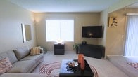 This single-floor residence features a well-appointed living area designed for both comfort and functionality. The living room is anchored by soft, wall-to-wall carpeting in a neutral tone that complements the warm beige walls throughout. A large window fitted with horizontal blinds allows natural light to fill the space, creating a bright and welcoming atmosphere.

The room is thoughtfully arranged with a contemporary sofa accented by decorative pillows, positioned across from a wall-mounted flat-screen television supported by a sleek, low-profile media console finished in dark tones. A matching coffee table with a glass-top surface serves as a central focal point, styled with decorative accessories including a bowl of fruit and a tall orange accent vase, adding a pop of color to the neutral palette.

Additional furnishings include a compact dark side table and modest indoor greenery that introduces a natural element to the interior. A textured area rug with a circular pattern grounds the seating arrangement and adds visual interest to the carpeted floor. The textured ceiling and clean wall surfaces contribute to the overall streamlined aesthetic of the space.