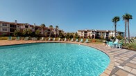 This inviting pool facility offers a seamless blend of outdoor relaxation and indoor recreation. The centerpiece is a generously sized outdoor swimming pool with crystal-clear blue water, surrounded by a wide tiled deck lined with lounge chairs — perfect for soaking up the sun against a backdrop of lush palm trees and a multi-story residential building. The pool area is enclosed by a wrought-iron fence, providing both security and an open, airy feel under clear skies.

The accompanying clubhouse interior is designed with a coastal aesthetic, featuring soft seafoam-blue walls, light tile flooring, and soaring ceilings with arched architectural details that lend an elegant, open atmosphere. Natural light pours in through large arched windows throughout.

Inside, a well-appointed kitchenette sits beneath a graceful archway, complete with gray cabinetry, granite countertops, a stainless steel refrigerator, and a microwave — ideal for entertaining. A rustic dining area with a concrete-top table and wicker chairs provides comfortable seating nearby.

The recreational spaces include a full-size pool table beneath a statement wrought-iron chandelier, a foosball table set on a patterned area rug, and a relaxed lounge nook with plush seating. French doors connect the interior directly to the pool deck, creating a fluid indoor-outdoor experience throughout the facility.
