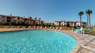 This inviting pool facility offers a seamless blend of outdoor relaxation and indoor recreation. The centerpiece is a generously sized outdoor swimming pool with crystal-clear blue water, surrounded by a wide tiled deck lined with lounge chairs — perfect for soaking up the sun against a backdrop of lush palm trees and a multi-story residential building. The pool area is enclosed by a wrought-iron fence, providing both security and an open, airy feel under clear skies.

The accompanying clubhouse interior is designed with a coastal aesthetic, featuring soft seafoam-blue walls, light tile flooring, and soaring ceilings with arched architectural details that lend an elegant, open atmosphere. Natural light pours in through large arched windows throughout.

Inside, a well-appointed kitchenette sits beneath a graceful archway, complete with gray cabinetry, granite countertops, a stainless steel refrigerator, and a microwave — ideal for entertaining. A rustic dining area with a concrete-top table and wicker chairs provides comfortable seating nearby.

The recreational spaces include a full-size pool table beneath a statement wrought-iron chandelier, a foosball table set on a patterned area rug, and a relaxed lounge nook with plush seating. French doors connect the interior directly to the pool deck, creating a fluid indoor-outdoor experience throughout the facility.