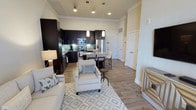 This single-floor residence showcases a thoughtfully designed open-concept layout that seamlessly connects the living, dining, and kitchen areas into one cohesive space. The living area is anchored by light-toned, neutral furnishings, including a plush sofa and accent chair arranged over a geometric-patterned area rug, creating a welcoming atmosphere. Warm wood-look flooring extends throughout, adding consistency and a sense of continuity across the space.

The kitchen is a standout feature, finished with dark espresso cabinetry that contrasts sharply against light countertops and a subtle tile backsplash. Stainless steel appliances, including a refrigerator, microwave, and range, provide a polished, modern aesthetic. Pendant lighting above the kitchen island adds both functional illumination and a contemporary design touch. The island doubles as a dining space, accommodating bar-height seating for casual meals.

Crown molding runs along the upper walls, adding a refined architectural detail. Recessed lighting throughout ensures even illumination across the open floor plan. Multiple doorways suggest additional private rooms beyond the main living area. Overall, the space balances modern finishes with a clean, neutral palette to create a functional and inviting environment.