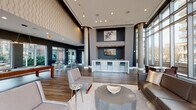 This contemporary amenity lounge is defined by its dramatic architectural scale, with soaring double-height ceilings and expansive floor-to-ceiling glazing that floods the interior with natural light and frames views of the surrounding urban streetscape. Exposed concrete columns punctuate the open-plan layout, lending an industrial edge to the otherwise polished interior. Hardwood-style flooring runs throughout, unifying the distinct activity zones within the space.

The lounge area serves as the social centerpiece, featuring a generous sectional sofa, accent chairs, and a substantial stone coffee table arranged atop a textured area rug. A centrally located wet bar with marble-style countertops, stainless steel appliances, a sink, and a wall-mounted television provides a functional entertaining hub. Decorative artwork and a textured accent wall add visual depth near the bar.

A billiards table occupies a dedicated game zone, while high-top bar tables with modern seating offer casual gathering spots near the windows. Lush potted palms and tropical plants are strategically placed throughout, softening the contemporary palette of dark navy, charcoal, and warm wood tones. The overall ambiance is polished, welcoming, and well-suited for social interaction.