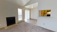 Located at 5741 Oscan Rd in Albuquerque, New Mexico, this single-story home offers a functional and comfortable layout with well-defined living spaces. The living room serves as the heart of the home, featuring plush carpeting and a tile-surround fireplace that provides a warm focal point. Natural light pours in through a large side window, brightening the space throughout the day. A sliding glass door with vertical blinds opens to the outdoors, offering convenient exterior access and additional light. The transition from carpet to tile flooring marks the shift from the living area into an adjoining space, creating a natural flow between zones. A pass-through window connects the living area to the kitchen, adding an open, communicative feel between rooms and allowing views of the wood-toned cabinetry beyond. The home's neutral color palette of soft whites and warm beige tones throughout the walls and flooring contributes to a clean, inviting ambiance. The angled ceiling detail in the living room adds a subtle architectural dimension to the otherwise streamlined interior. Overall, the home presents a practical and well-maintained single-floor layout suited to comfortable everyday living in the Albuquerque area.
