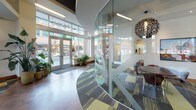 Located at 2 E Main St in downtown Auburn, Washington, this single-floor commercial lobby space offers a thoughtfully designed environment that blends modern aesthetics with welcoming functionality. The entrance features floor-to-ceiling glass doors and expansive windows that flood the interior with abundant natural light while providing clear views of the surrounding streetscape.

The main lounge area is anchored by a sweeping curved sectional sofa in charcoal gray, accented with vibrant throw pillows in yellow, red, and teal. Striking sculptural pendant lights hang above, adding a dramatic focal point to the seating area. Patterned area rugs in yellow, gray, and charcoal tones complement the overall color palette and define distinct zones within the open layout.

A kitchenette area features sleek white cabinetry with green upper cabinets, concrete-look wall panels, dark wood accents, polished countertops, and a built-in coffee station — creating a functional yet stylish refreshment nook. The flooring transitions between polished concrete and patterned carpet, further distinguishing the various areas.

Large indoor plants are strategically placed throughout, adding organic warmth to the contemporary design. Curated urban photography adorns the walls, reinforcing a connection to the surrounding community. An elevator alcove with textured stone walls and a glass-partitioned conference area complete this well-appointed, light-filled commercial space.