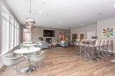 This single-floor community space in Statesboro, Georgia, is designed with a modern aesthetic that balances comfort and functionality. The main lounge area is anchored by a white-painted brick fireplace with a large wall-mounted television above it, creating a warm, inviting focal point. Plush gray sofas and area rugs are thoughtfully arranged around the hearth, while vibrant, colorful abstract artwork adds visual energy throughout the space. Wood-look flooring flows seamlessly across the entire floor plan, contributing to a cohesive and welcoming atmosphere.

Large, floor-to-ceiling windows line one side of the lounge, flooding the interior with abundant natural light and offering direct views of an outdoor pool area. Pendant lighting in metallic finishes adds a contemporary touch overhead. Scattered round tables with modern molded chairs provide flexible seating for small groups, making the space suitable for both socializing and casual work.

A well-appointed kitchenette features gray shaker-style cabinetry, light quartz countertops, a deep undermount sink, a beverage refrigerator, and a bold navy subway tile backsplash with floating wood shelves above. A high-top table with bar-height seating nearby offers an additional casual dining option. The overall ambiance is bright, modern, and thoughtfully curated.