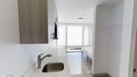 This efficiently designed studio with one bathroom makes smart use of its compact footprint. The open-plan layout integrates the living and sleeping areas into a single cohesive space, finished with light wood-tone flooring that adds warmth throughout.

The kitchenette is outfitted with contemporary dark wood cabinetry, a quartz-style countertop, and a stainless steel sink with a brushed nickel faucet. Upper cabinetry provides practical storage, while the clean lines and neutral palette keep the space feeling open and uncluttered.

A wall-mounted television is positioned conveniently between the kitchen and living area, maximizing functionality without sacrificing floor space. At the far end of the unit, a large multi-pane window allows abundant natural light to pour in, brightening the sleeping area and offering a view of the surrounding environment. The white walls and bright ceiling further enhance the sense of openness.

Ceiling-mounted lighting provides even illumination throughout the unit. The overall design aesthetic is clean and modern, with a neutral color scheme of whites, grays, and warm wood tones that creates a cohesive and livable environment. Despite its compact size, the thoughtful layout and bright finishes make this studio feel functional and comfortable.