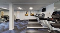 Located at 1037 Southern Artery in Quincy, MA, this single-level fitness center offers a well-organized workout space designed to accommodate a variety of exercise routines. The room features a drop ceiling with recessed fluorescent lighting that ensures consistent, even illumination throughout the space.

The cardio area is prominently outfitted with multiple treadmills arranged in a row, complemented by an elliptical trainer and a stationary bike, providing users with diverse aerobic workout options. A wall-mounted flat-screen television is positioned for easy viewing during cardio sessions. Motivational framed artwork depicting athletic silhouettes against warm golden backgrounds accents the white walls, adding visual energy to the space.

A dedicated strength training section houses a tiered dumbbell rack with a full range of free weights, reflected in a large wall mirror that visually expands the room. Additional resistance and stretching equipment rounds out the training options available.

The flooring throughout consists of patterned carpet tiles in a chevron design, blending blue and grey tones for a modern aesthetic. Windows along one wall allow natural light to enter and offer views of the surrounding greenery, enhancing the overall atmosphere of the space.