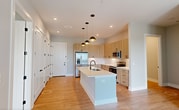 This 1,110-square-foot single-floor residence at 4362 Watermark Dr in Wilmington, NC features a clean, modern design with a neutral color palette throughout. The open-concept kitchen and living area serves as the heart of the home, showcasing light wood cabinetry, a white quartz island with an undermount sink, stainless steel appliances including a French door refrigerator and range, a subway tile backsplash, and stylish pendant lighting. Light wood-tone flooring runs through the main living areas, adding warmth to the contemporary aesthetic. The primary bedroom spans 190 square feet with direct balcony access via a glass-paned door, while the second bedroom features plush carpeting, a ceiling fan, and a large window with open views. Both bedrooms are accompanied by dedicated closets, including a walk-in closet with built-in shelving and chrome hanging rods. The primary bathroom includes a glass-enclosed shower with black hardware and direct access to the walk-in closet. The private balcony features modern black metal railing and offers sweeping views of a scenic waterway and open marsh, characteristic of Wilmington's coastal landscape. The building exterior features contemporary horizontal gray siding with clean architectural lines.