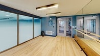 Located at 18750 Delaware St in Huntington Beach, California, this dedicated fitness studio offers a well-appointed, single-room exercise space designed for group and individual training. The room features light hardwood flooring that lends a clean, contemporary feel throughout. A striking full-width frosted glass partition wall with dark bronze framing separates the studio from an adjacent space, allowing diffused light to filter through while maintaining privacy.

The studio is equipped with spin bikes positioned for class-style cycling sessions, along with an assortment of fitness accessories including stability balls, resistance bands, and yoga mats stored along the walls. A large wall-mounted display screen serves as a focal point for guided workout sessions and connected fitness programming. A full-length mirror along one wall enhances the sense of space and supports functional training.

The walls are finished in a bold navy and teal blue palette, adding an energetic ambiance suited for an active environment. Recessed and linear LED lighting along the ceiling provides bright, even illumination throughout. The studio is accessed through a glass-paneled door, and ventilation is supported by integrated HVAC registers. Overall, the space presents a modern, purpose-built fitness environment well-suited for cycling classes, yoga, and cross-training activities.