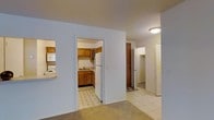 This single-floor residential space offers a practical and well-organized layout designed for comfortable everyday living. The kitchen is a standout functional area, outfitted with warm wood cabinetry providing ample storage, a white refrigerator, a range with overhead microwave, and a sink set beneath the cabinetry. Light-toned tile flooring in the kitchen adds a clean, easy-to-maintain surface. A pass-through window connects the kitchen to the adjacent living area, enhancing the sense of openness between spaces. The main living and hallway areas feature neutral carpeting and light-toned tile, creating a cohesive and understated aesthetic throughout. Smooth white walls and flush ceiling lighting keep the interior bright and uncluttered. The hallway leads to additional areas of the unit, with a louvered closet door visible along the corridor, offering convenient built-in storage. A secondary doorway, framed in wood tones, provides access to further rooms within the space. The overall ambiance is clean, simple, and move-in ready, making it well-suited for those seeking a straightforward, functional living environment with all essential spaces thoughtfully arranged on a single level.