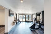 Located at 600 Antlers Lane in Charlotte, NC, this well-appointed fitness center offers a dedicated exercise space designed to support a full range of workout routines. The room features sleek hardwood-style plank flooring in cool grey tones, crisp white and grey accent walls, and recessed lighting that creates a bright, energizing atmosphere.

The cardio zone is equipped with multiple TRUE-brand treadmills, a recumbent bike, an upright stationary bike, and a rowing-style machine, providing a comprehensive selection of aerobic options. A dumbbell rack and adjustable weight bench round out the strength training area. A wall-mounted flat-screen television is positioned for easy viewing during workouts, and a water fountain station is conveniently accessible within the space.

Natural light flows in through multiple windows and a set of glass-paned French doors that open directly toward an outdoor pool area, offering refreshing views of the surrounding greenery and community buildings. Motivational fitness artwork adorns the walls, contributing to an upbeat and encouraging environment. The space is thoughtfully laid out to allow for comfortable movement between equipment, making it a practical and inviting facility for everyday fitness needs.