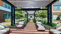 This single-floor space combines functional interior storage with access to an impressive outdoor amenity area. The interior is finished with neutral, cream-toned walls and classic two-panel doors fitted with sleek brushed nickel hardware, lending a clean and timeless aesthetic throughout.

A notable interior feature is the walk-in closet, which offers generous storage capacity. It is outfitted with a comprehensive white wire shelving system, including multiple stacked shelving units along one wall and a dedicated hanging rod with an upper storage shelf along another. Neutral carpeting covers the closet floor, adding a soft, practical finish to the space.

The outdoor courtyard is a standout amenity. A structured pergola with dark metal posts and wooden overhead slats runs the length of a herringbone-patterned brick walkway, draped with string lights for evening ambiance. Wicker lounge chairs with plush white cushions are arranged beneath the pergola, creating comfortable seating nodes surrounded by mature trees, flowering shrubs, and manicured garden beds. The courtyard faces a multi-story brick and panel building facade with balconies and large windows, reinforcing a well-designed residential community setting.