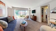 This single-floor residence offers a thoughtfully designed layout that balances comfort and functionality across multiple distinct living spaces. The living room is generously sized, featuring plush wall-to-wall carpeting, neutral walls, and a large sliding glass door that fills the room with natural light and provides direct access to an outdoor patio with views of lush greenery. A built-in shelving nook adds practical storage near the living area.

The separate dining room accommodates a full dining set beneath a pendant light fixture, with a window dressed in floor-length curtains that allows natural light to brighten the space. The kitchen is equipped with warm oak cabinetry, dark granite-style countertops, hardwood-style flooring, and black appliances including a gas range, dishwasher, and range hood.

The primary bedroom is spacious and carpeted, offering ample room for a king-sized bed and generous dresser storage. A second bedroom features soft carpet flooring, a large window with natural light, and a clean, restful ambiance. The full bathroom includes hardwood-style flooring, a tub-shower combination, a vanity, and bright, clean finishes throughout. The overall design of the residence strikes a balance between mid-century modern influences and everyday practicality.