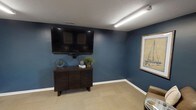 This single-floor space presents a thoughtfully designed lounge or sitting area that combines comfort with a cohesive aesthetic. The most prominent feature is the rich, deep blue wall color, which lends the room a calm yet bold character. A large wall-mounted flat-screen television serves as a central focal point, positioned above a dark-stained wooden media console with slatted panel detailing and mid-century modern legs. The console is accessorized with decorative items including a potted plant, small sculptures, and a round accent piece, adding warmth and personality to the space.

The flooring consists of neutral-toned, low-pile carpet in a subtle grid pattern, providing a clean contrast to the vibrant walls. White baseboards frame the room, offering a crisp visual boundary. A framed nautical-themed artwork adorns one wall, reinforcing a relaxed, coastal-inspired ambiance. Overhead, a linear LED light fixture provides bright, even illumination, complemented by a textured ceiling finish. A partial view of additional seating is visible at the edge of the frame, suggesting a comfortable and functional gathering area. Overall, the space communicates a polished, inviting atmosphere suited for relaxation or casual entertainment.